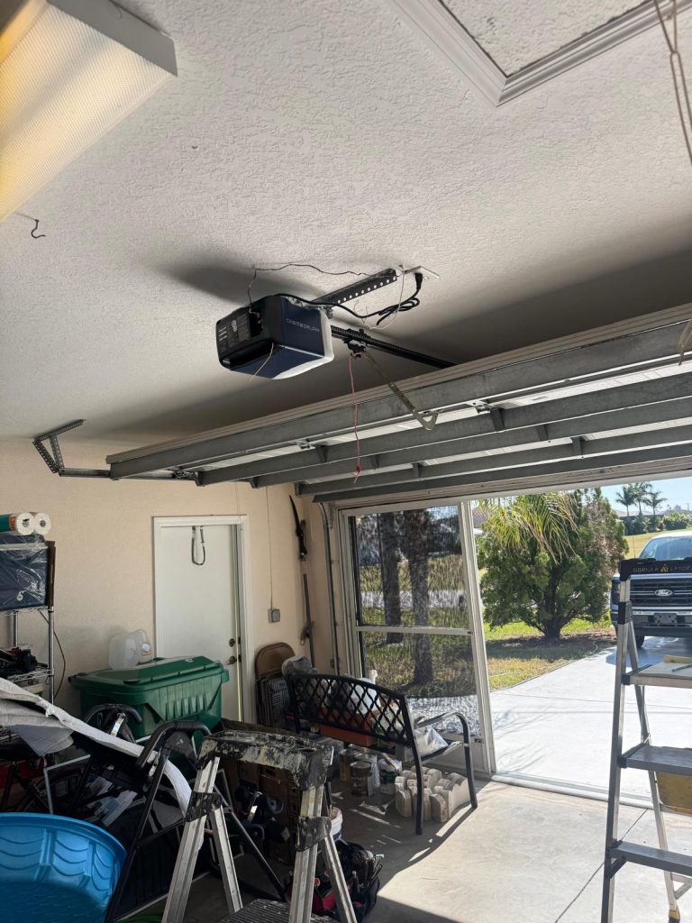 Garage with overhead garage door opener, cluttered workspace, and open door revealing exterior landscaping, illustrating the importance of garage door maintenance and cable replacement services.